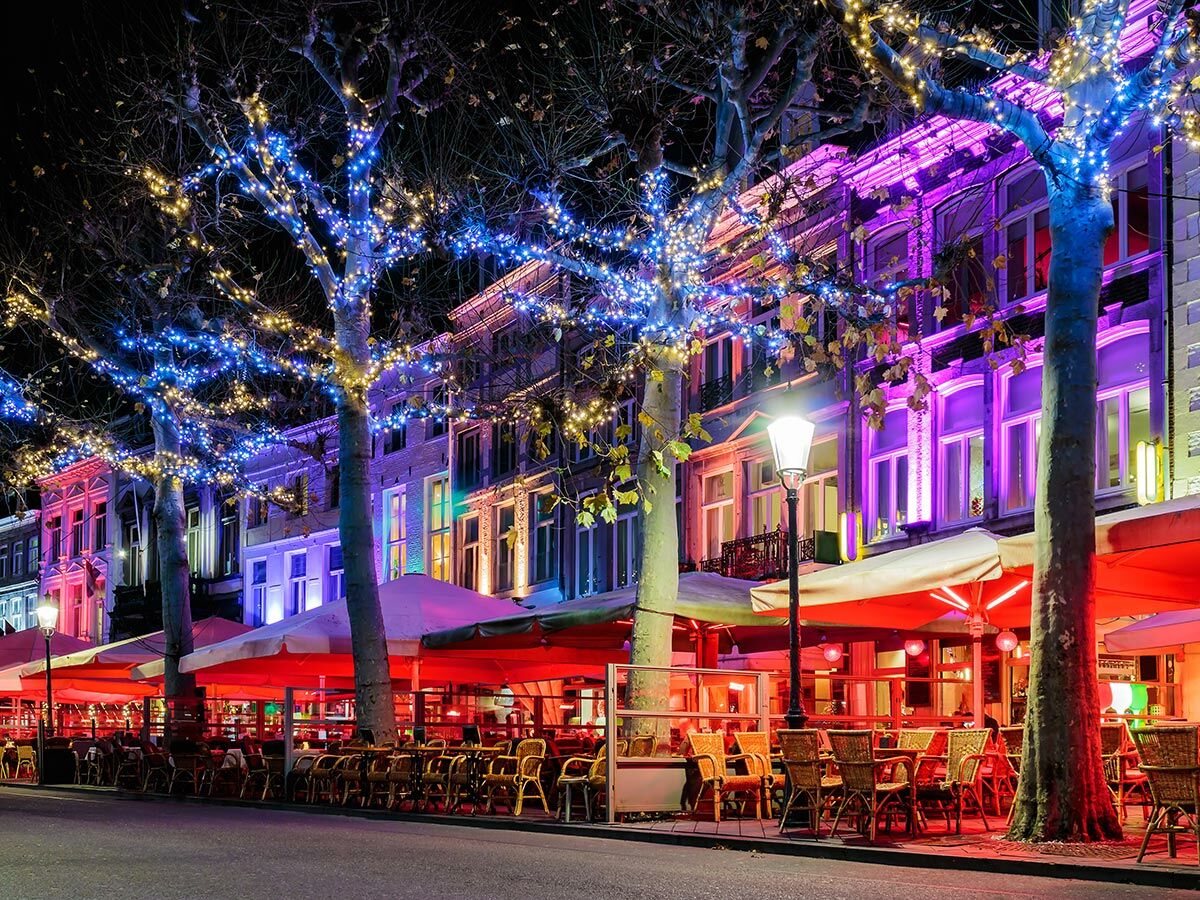Beleuchtete Stadtstraße bei Nacht. Bäume mit Lichterketten, bunte Gebäude und Straßencafés mit roten Sonnenschirmen und Sitzplätzen.