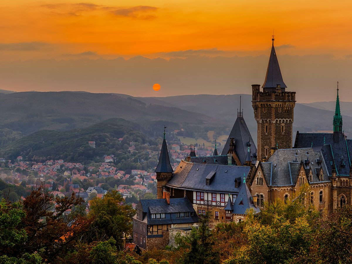 Märchenhaftes Schloss Wernigerode im Harz bei Sonnenuntergang über einem Dorf mit Bergen und orangefarbenem Himmel.