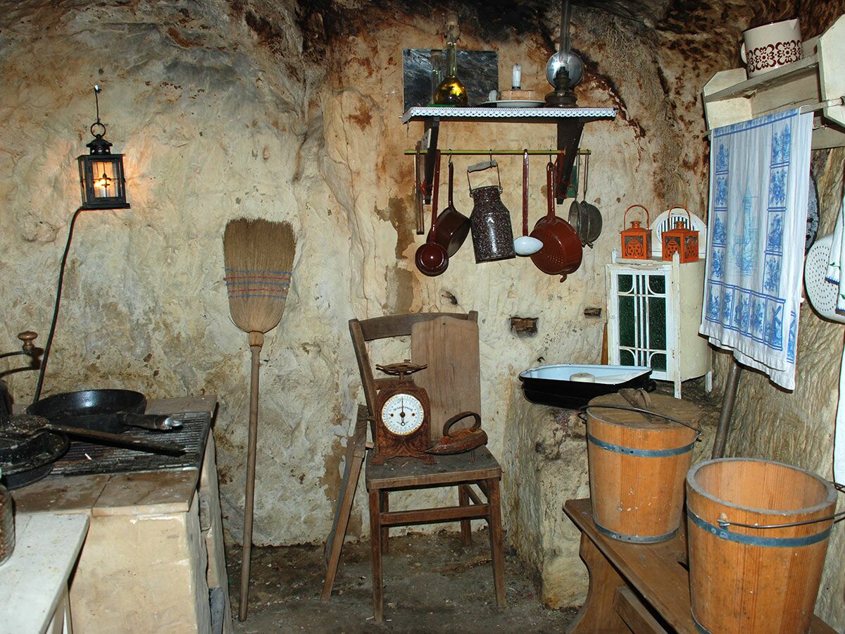 Rustikale Höhlenküche mit alten Küchenutensilien, Besen und Holzstühlen, beleuchtet von einer Wandlaterne.
