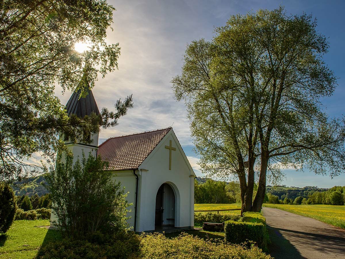 Kleine weiße Kapelle mit spitzem Dach in ländlicher Umgebung, umgeben von grünen Bäumen und Wiesen unter strahlendem Sonnenlicht.