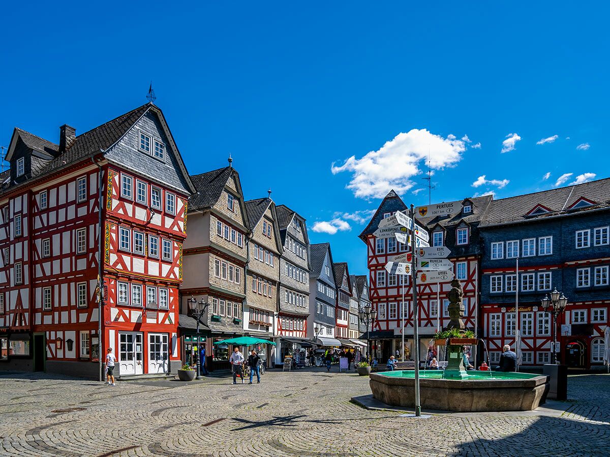 Bunter historischer Marktplatz mit Fachwerkhäusern, Brunnen und Menschen unter blauem Himmel in deutscher Altstadt.
