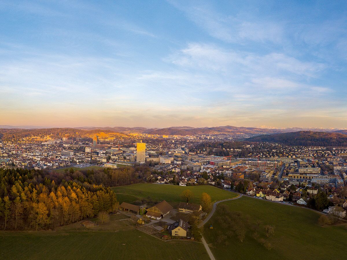 Luftaufnahme einer Stadt bei Sonnenuntergang mit Wald, Feldern, Gebäuden und schneebedeckten Alpen im Hintergrund.