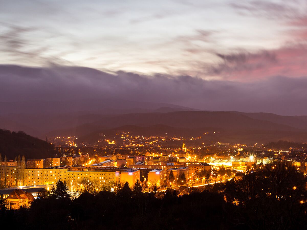 Panoramablick auf eine beleuchtete Stadt bei Einbruch der Dunkelheit, mit warmen Lichtern, Gebäuden und dunklen Bergen unter violettem Himmel.