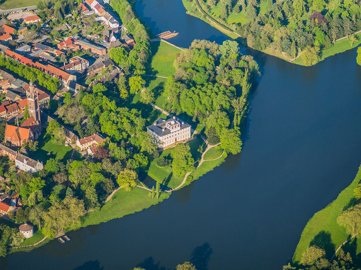 Luftaufnahme des Rheinsberger Schlosses, umgeben von grünen Bäumen und einem blauen See, daneben ein Dorf.