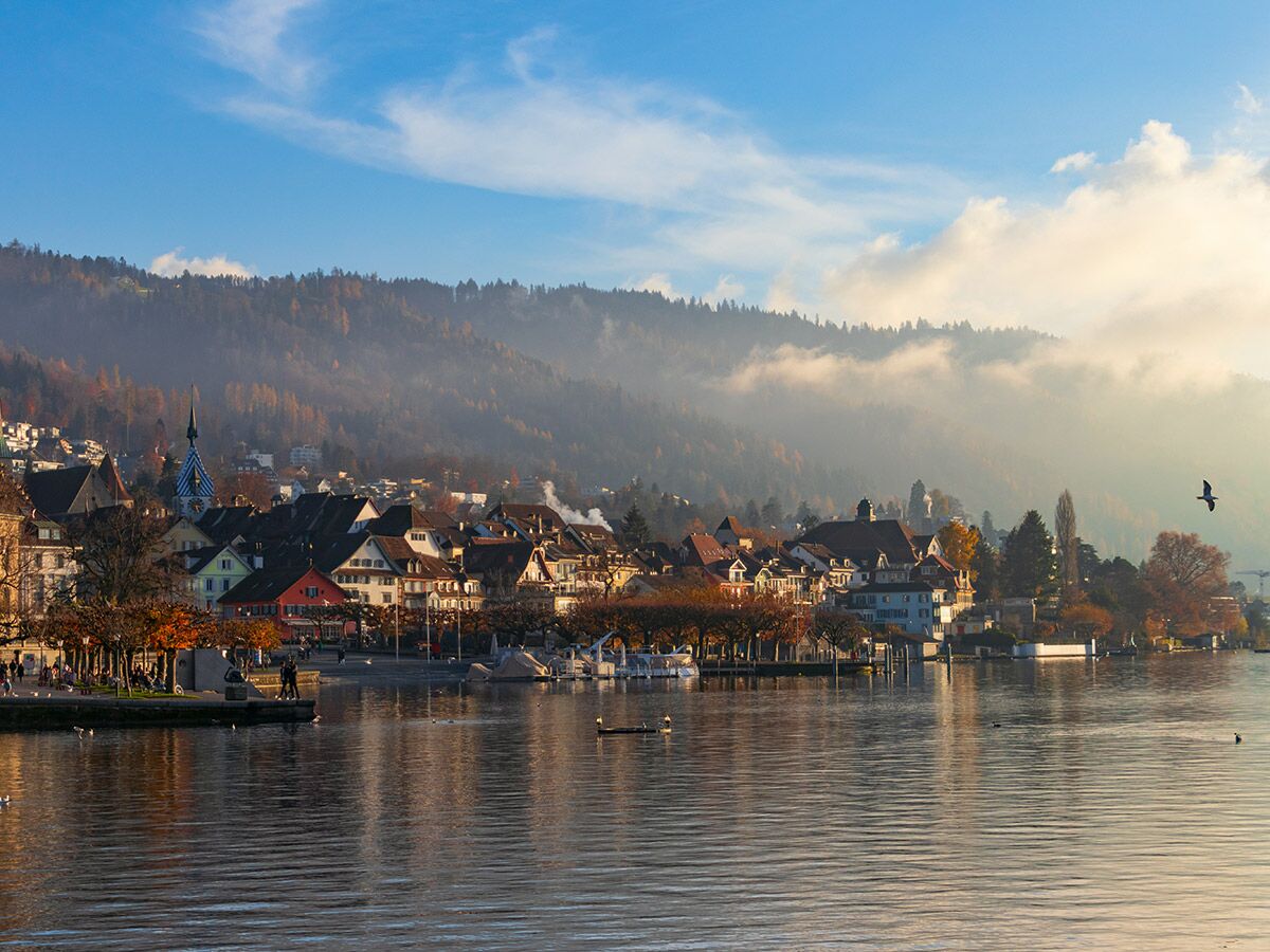 Panorama einer idyllischen Seestadt mit bunten Häusern, herbstlichen Bäumen, nebelverhangenen Bergen und einem Kirchturm bei Sonnenaufgang.