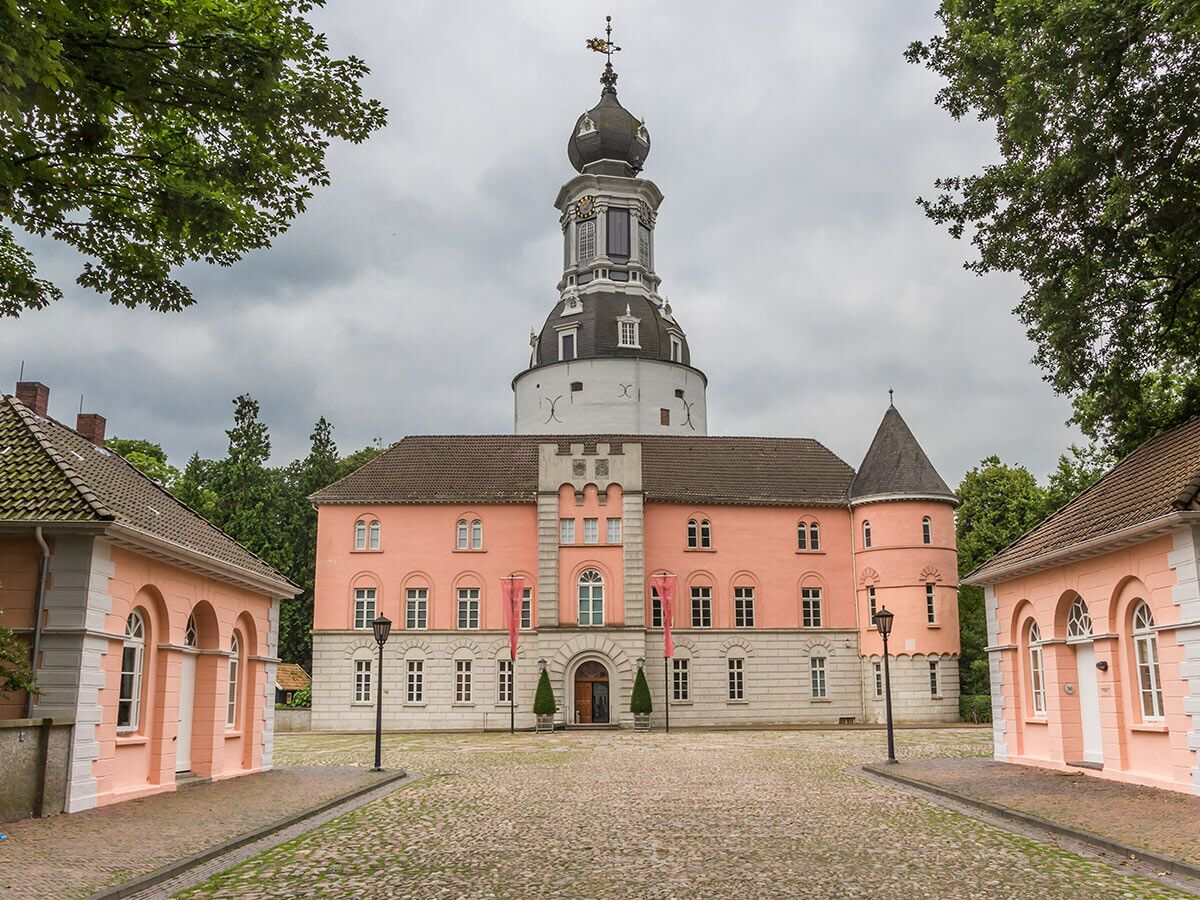 Barockes Schloss mit imposantem, weißem Turm und kuppelförmigem Dach, umgeben von rosa Gebäuden und Kopfsteinpflasterhof unter bewölktem Himmel.