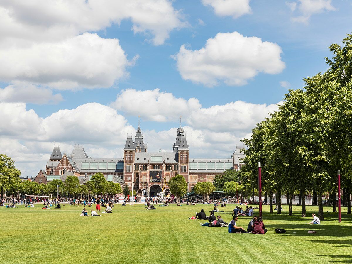 Menschen entspannen auf dem grünen Rasen des Museumpleins vor dem historischen Rijksmuseum in Amsterdam an einem sonnigen Tag.