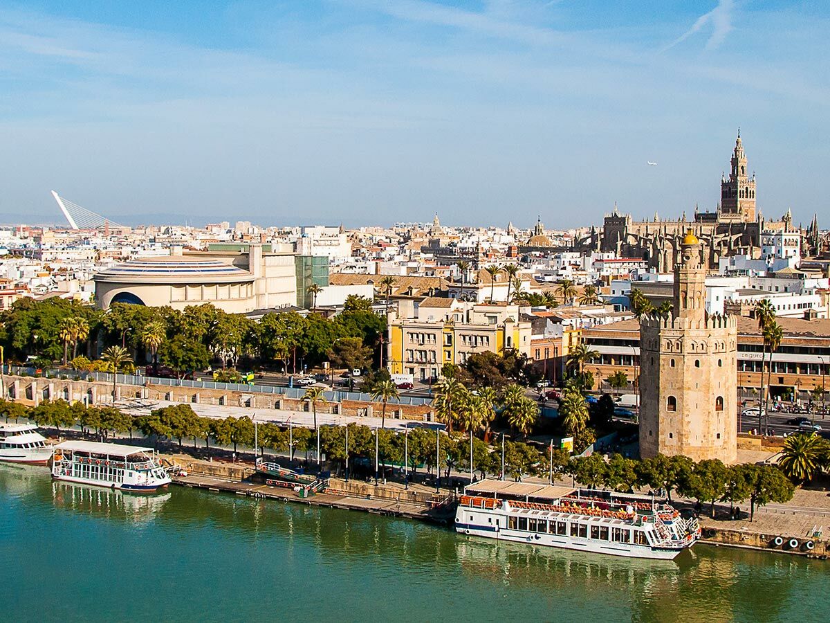 Panoramablick auf Sevilla mit dem Fluss Guadalquivir, Torre del Oro, der Kathedrale und Ausflugsbooten am Ufer bei Tag.