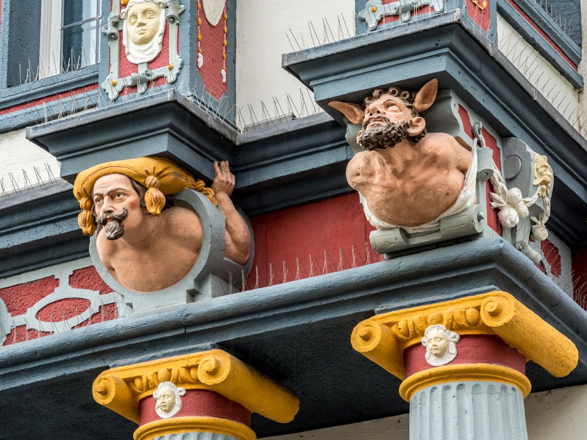 Detailansicht historischer Gebäude mit zwei farbenfrohen grotesken Skulpturen und Vogelschutzspitzen an der Fassade.