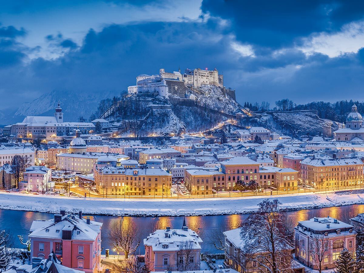 Panoramablick auf Salzburg im Winter bei Nacht. Die Hohensalzburg thront schneebedeckt über der beleuchteten Altstadt und der Salzach.