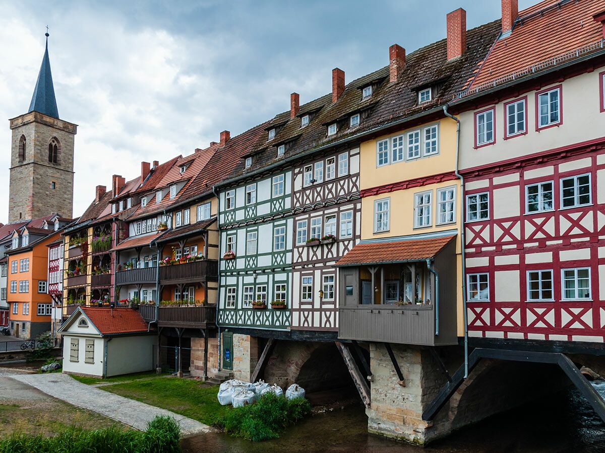 Bunte Fachwerkhäuser der Krämerbrücke in Erfurt, Deutschland, über Fluss mit Kirchturm unter bedecktem Himmel.
