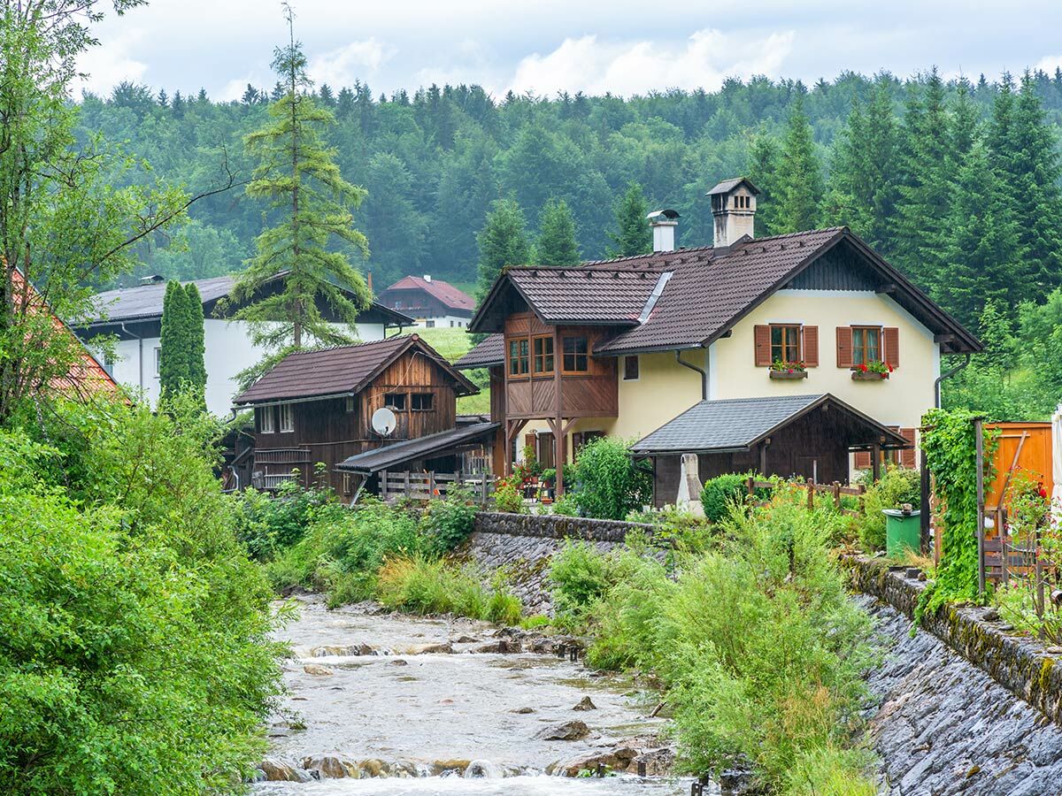 Dorf mit traditionellen Häusern am klaren Fluss, umgeben von dichtem Wald und grünen Hügeln. Idyllische Alpenlandschaft.