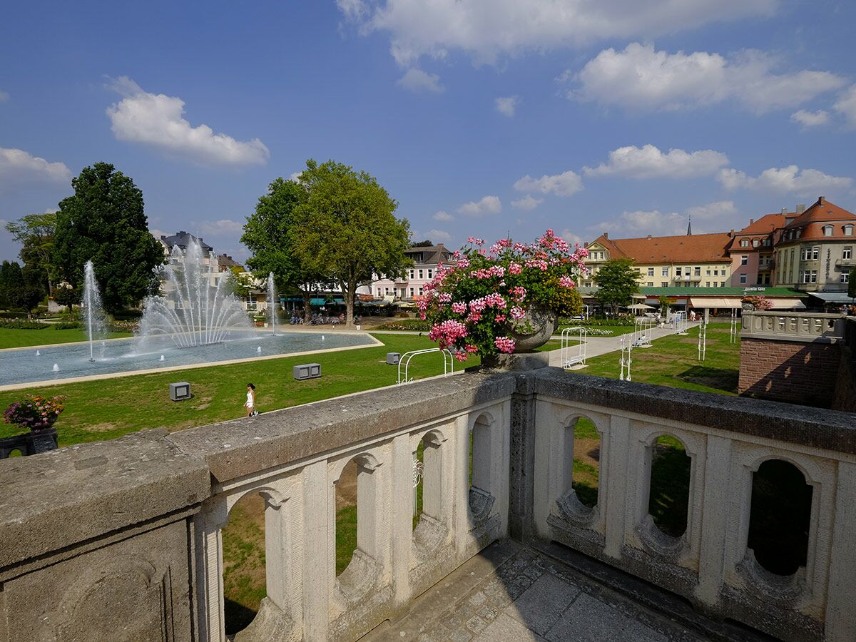 Sonniger Stadtpark mit Springbrunnen, üppigem Grün, pinken Geranien auf einer Balustrade und historischen Gebäuden im Hintergrund.