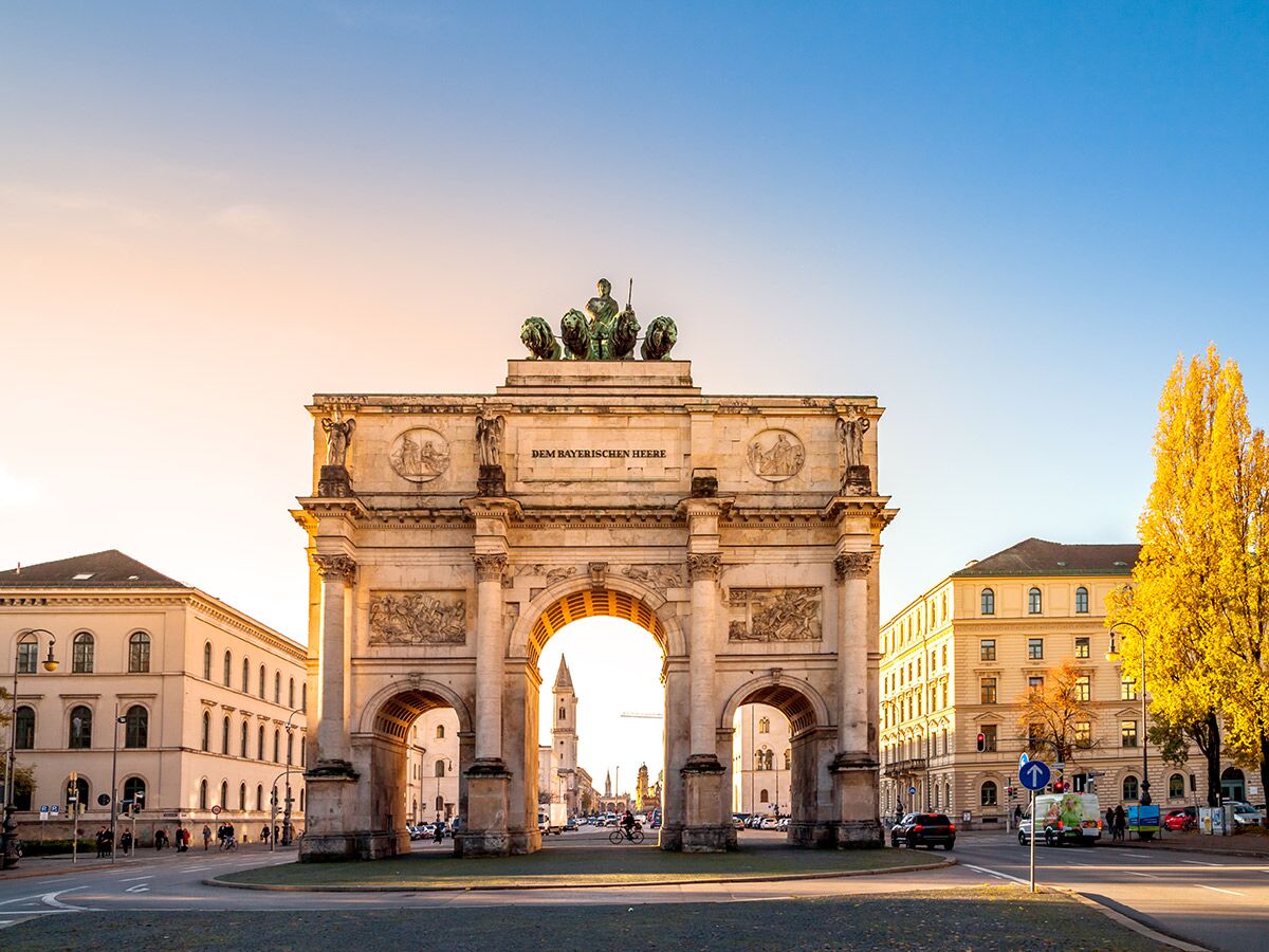 Das Siegestor in München bei goldenem Herbstlicht mit gelben Bäumen, Gebäuden und Straßenverkehr.