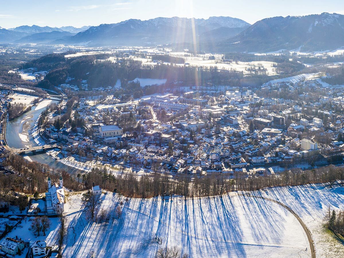 Luftaufnahme einer schneebedeckten Stadt im Tal mit Fluss, Brücken und einer Kirche, umgeben von sonnigen Alpenbergen.