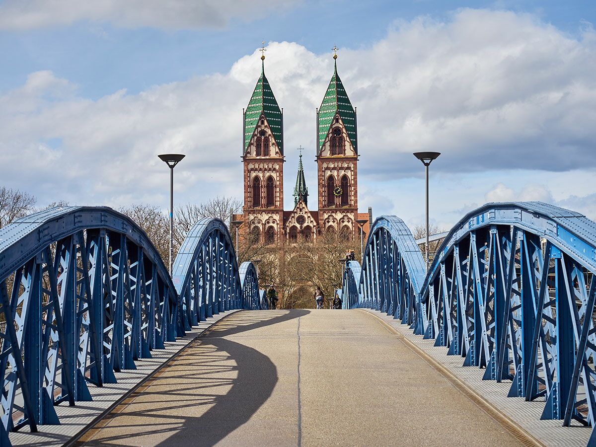Eine blaue Stahlbogenbrücke führt auf die Herz-Jesu-Kirche in Freiburg mit grünen Türmen und bewölktem Himmel zu.