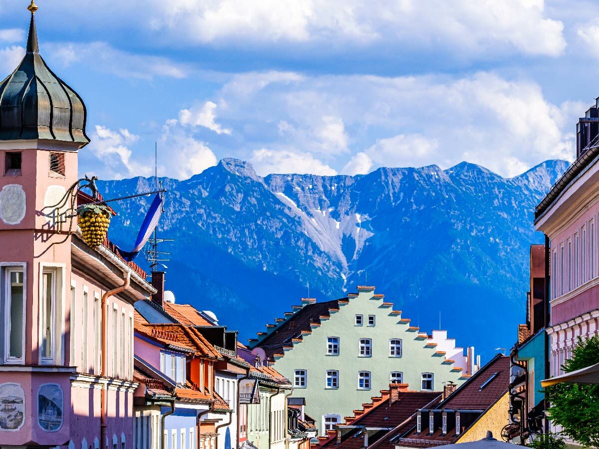 Bunte Gebäude einer malerischen Alpenstadt vor imposanten Bergen und blauem Himmel. Historische Architektur und Naturkulisse.
