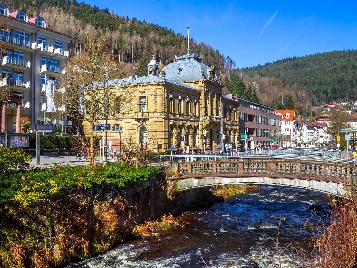 Bad Wildbad im Schwarzwald mit historischem Kurhaus, Enz-Fluss, Brücke und grünen Hügeln unter blauem Himmel.