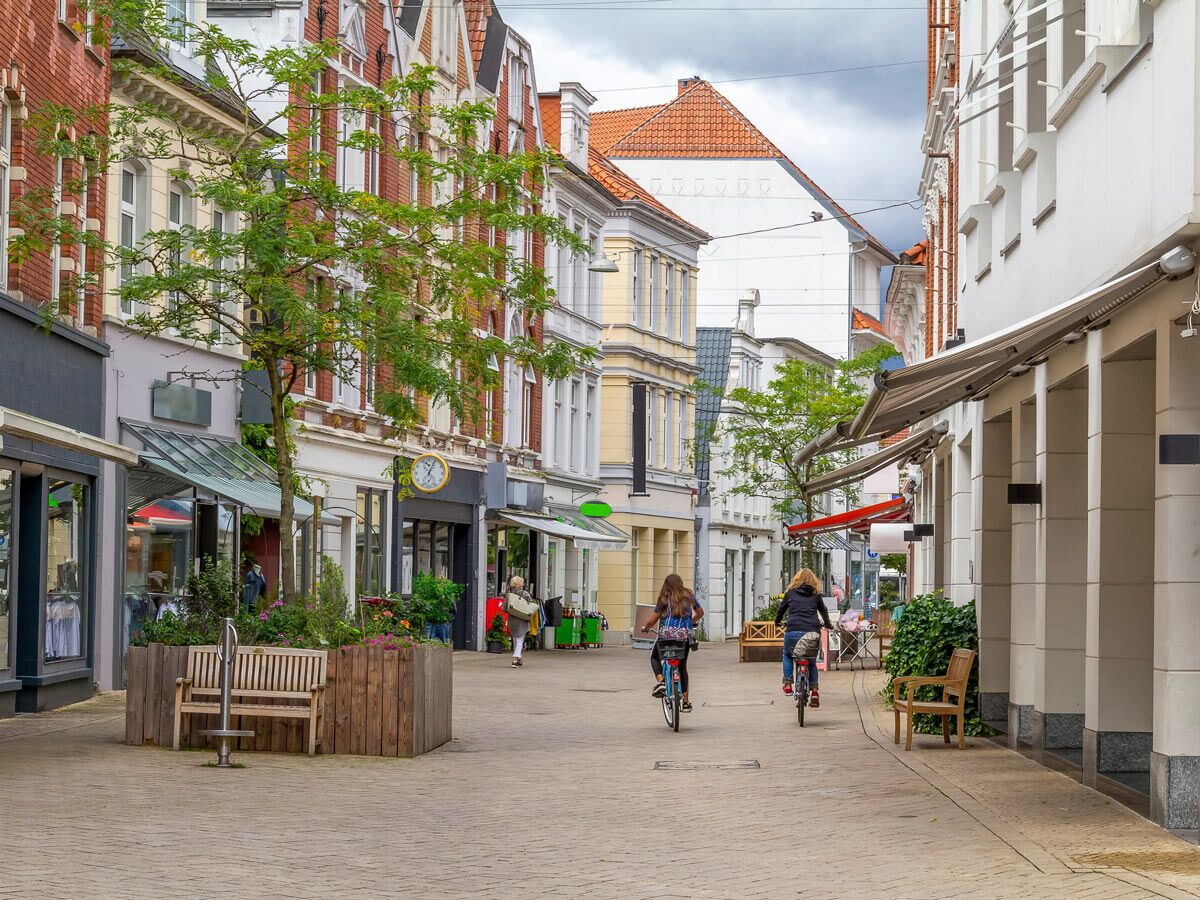 Belebte europäische Einkaufsstraße mit historischen Gebäuden, Geschäften, Bäumen, Bänken und zwei radelnden Frauen.