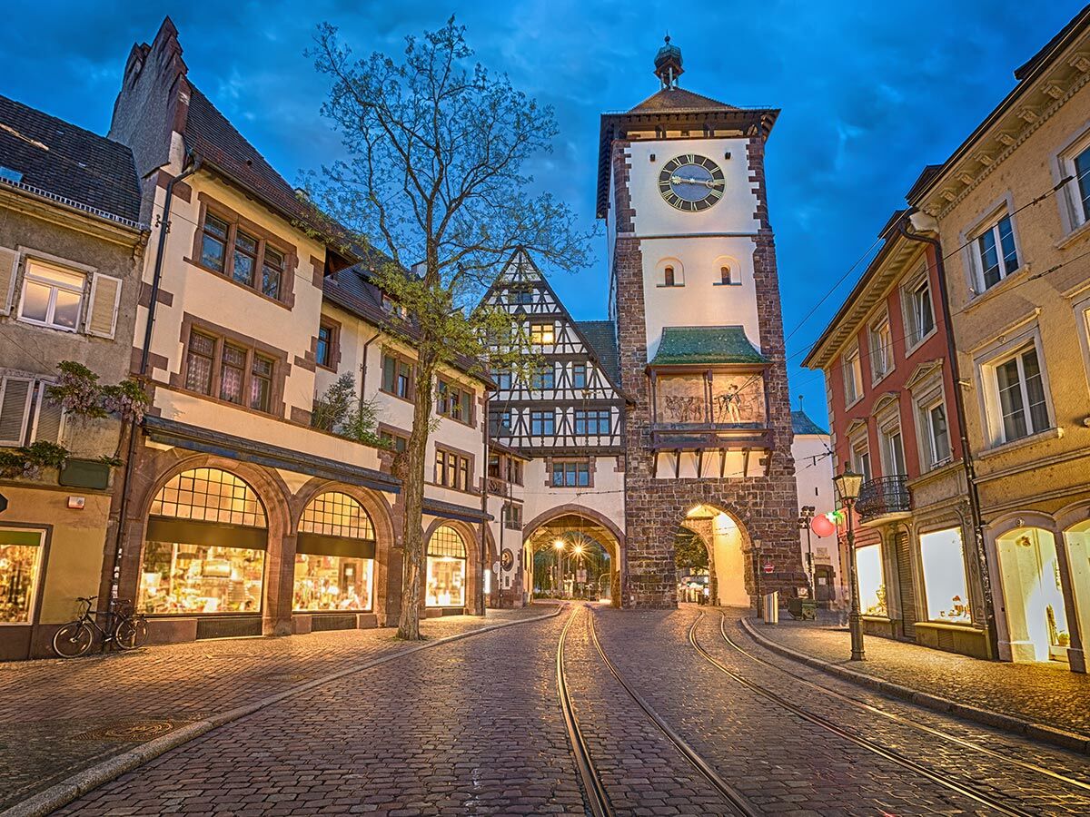 Beleuchtetes Martinstor in Freiburg im Breisgau bei Abenddämmerung mit Kopfsteinpflaster, Straßenbahnschienen und historischen Geschäften.