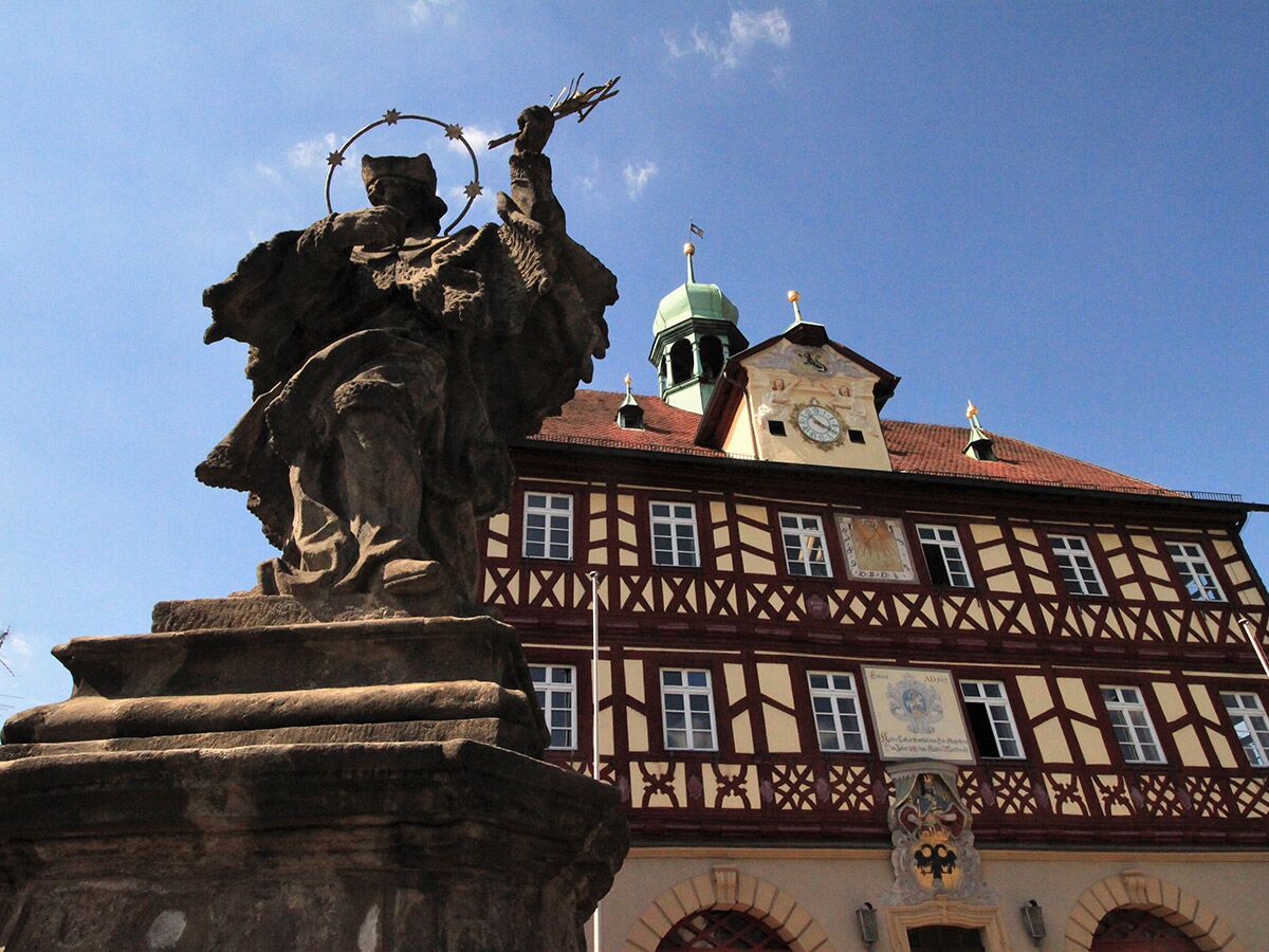 Große Steinstatue eines Heiligen vor einem historischen Fachwerk-Rathaus mit Uhrturm und Sonnenuhr bei blauem Himmel in Deutschland.