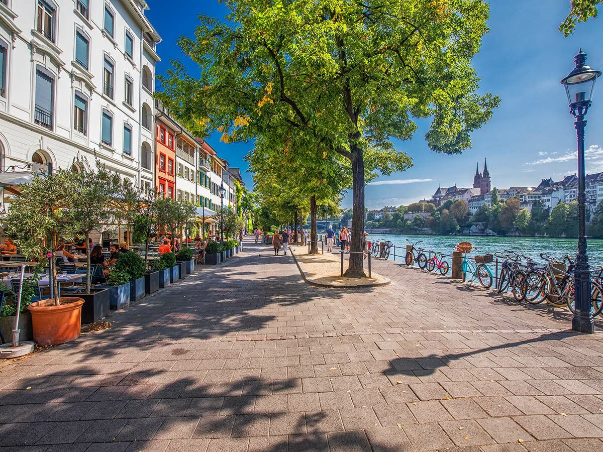 Blick auf die lebhafte Rheinpromenade in Basel mit Straßencafés, Bäumen, parkenden Fahrrädern, historischen Gebäuden und dem Basler Münster.