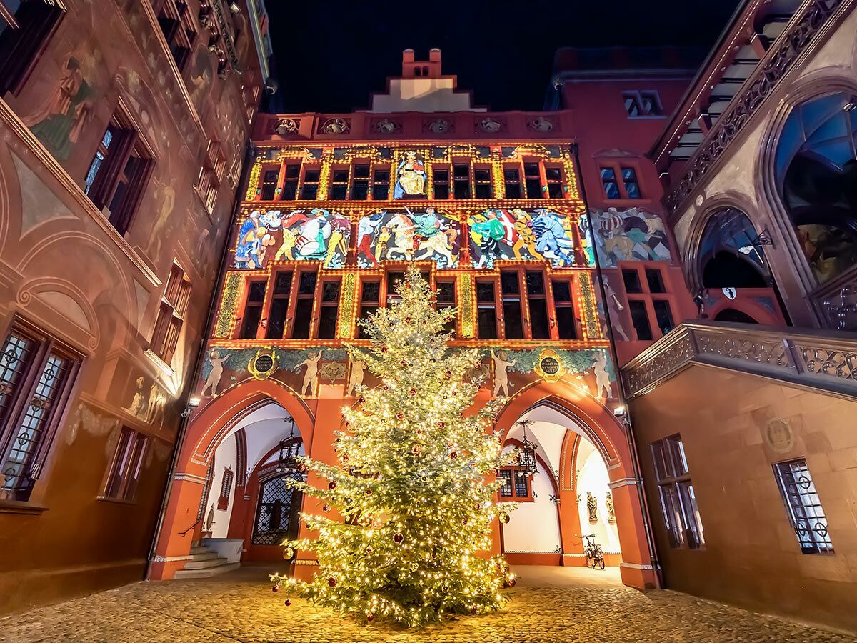 Festlich beleuchtetes Basler Rathaus mit großem, geschmücktem Weihnachtsbaum auf Kopfsteinpflasterplatz bei Nacht, farbenfrohe Fassadenprojektion und historische Architektur.