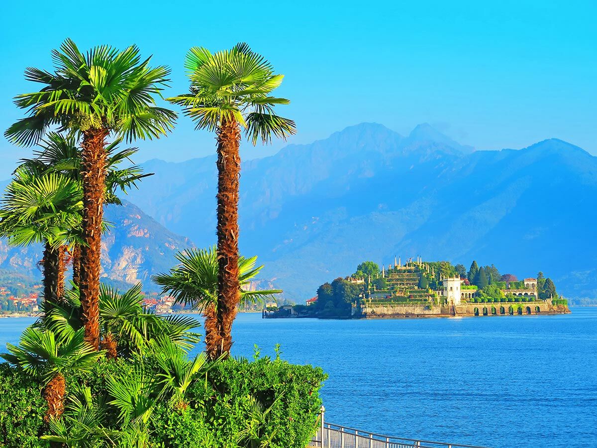 Panoramablick auf Isola Bella mit Palast, terrassierten Gärten und Palmen am Ufer des Lago Maggiore, umgeben von Bergen.