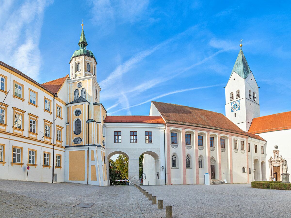 Historischer Klosterhof mit weiß-gelben Gebäuden, grünem Kuppelturm, hohem Kirchturm und Torbögen unter blauem Himmel.