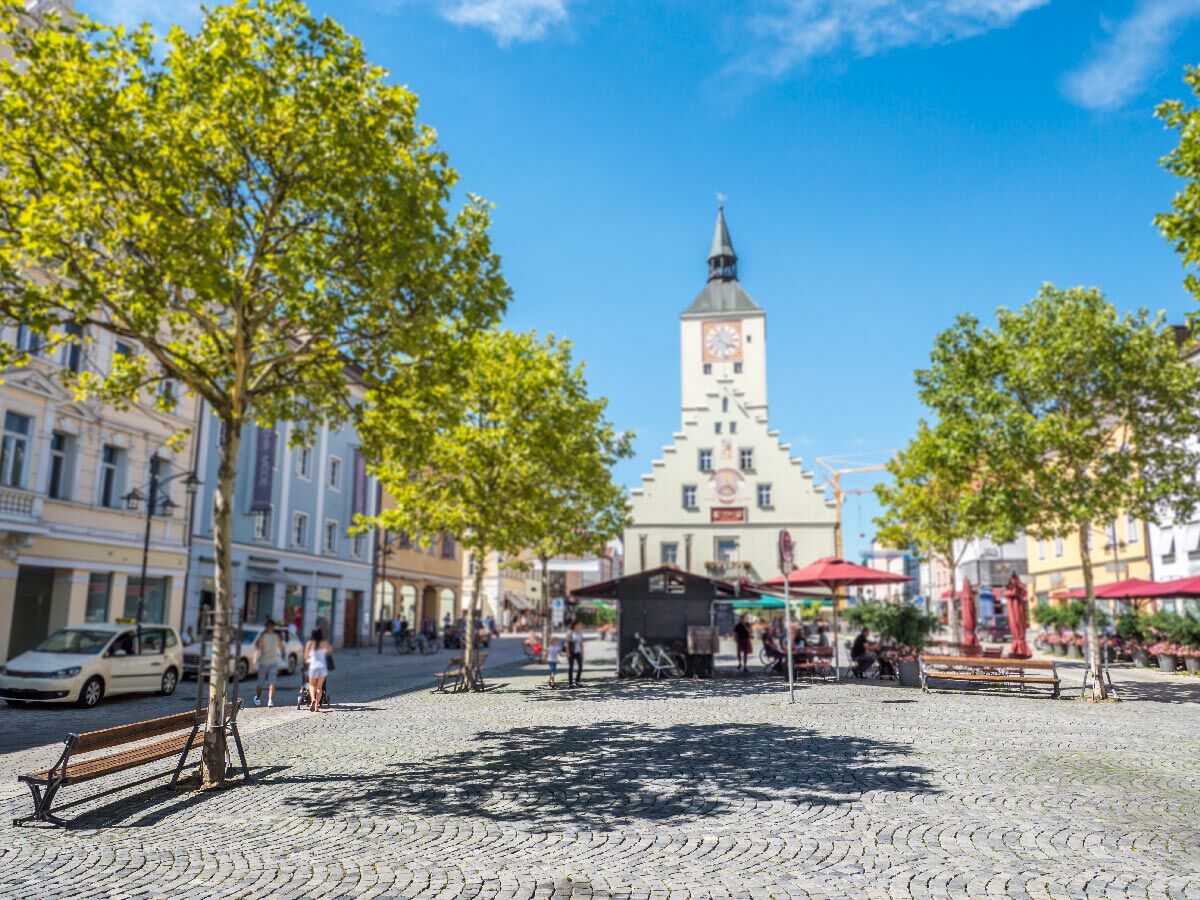 Historischer Marktplatz mit altem Rathaus und Uhr unter blauem Himmel, Bäume und Menschen genießen den sonnigen Tag.