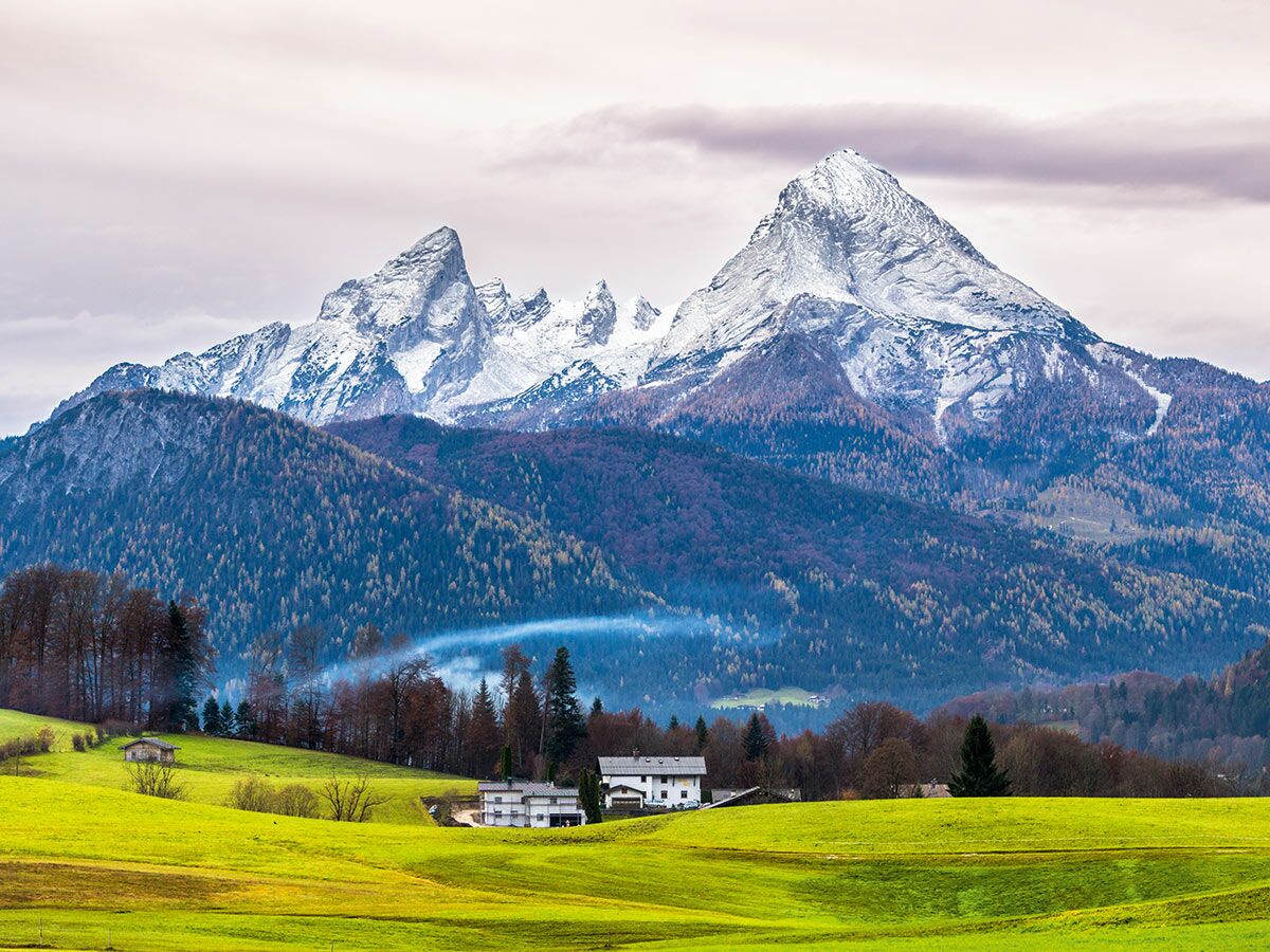 Malerische Alpenlandschaft mit schneebedeckten Gipfeln, dichten Wäldern, grünen Wiesen und kleinen Häusern unter bewölktem Himmel.