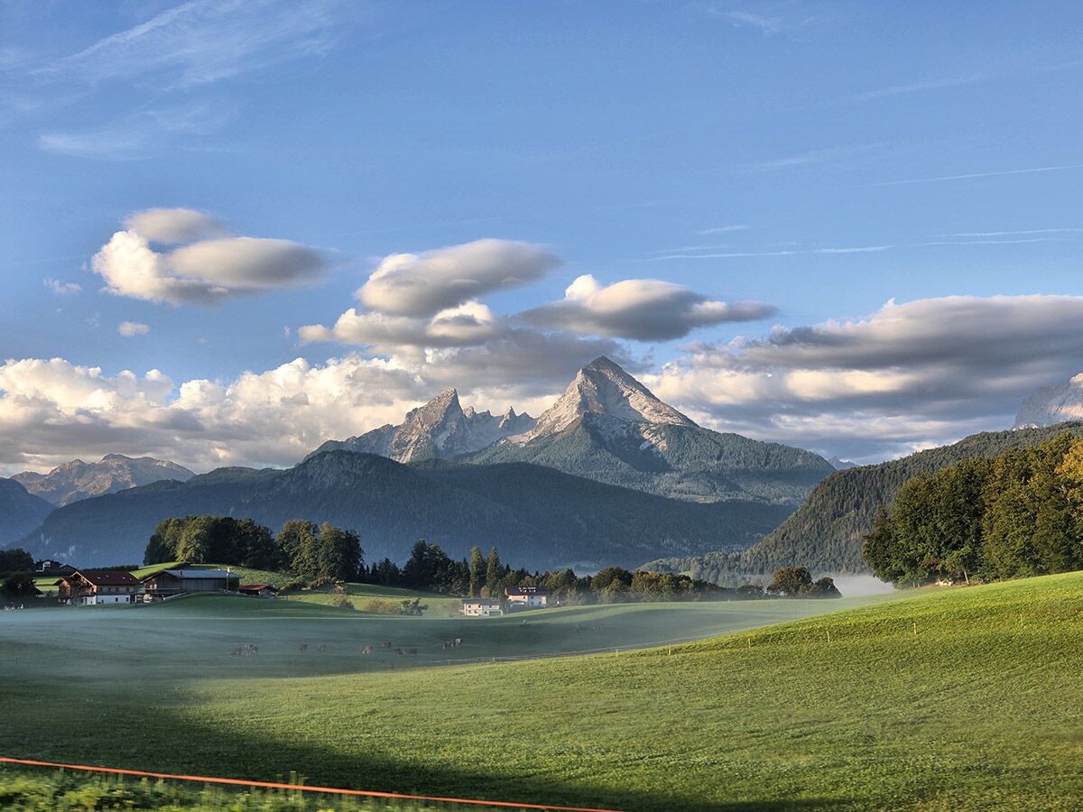 Idyllische Alpenlandschaft im Morgenlicht mit Watzmann-Massiv, grünen Wiesen, Bauernhöfen und Bodennebel. Kühe grasen friedlich.