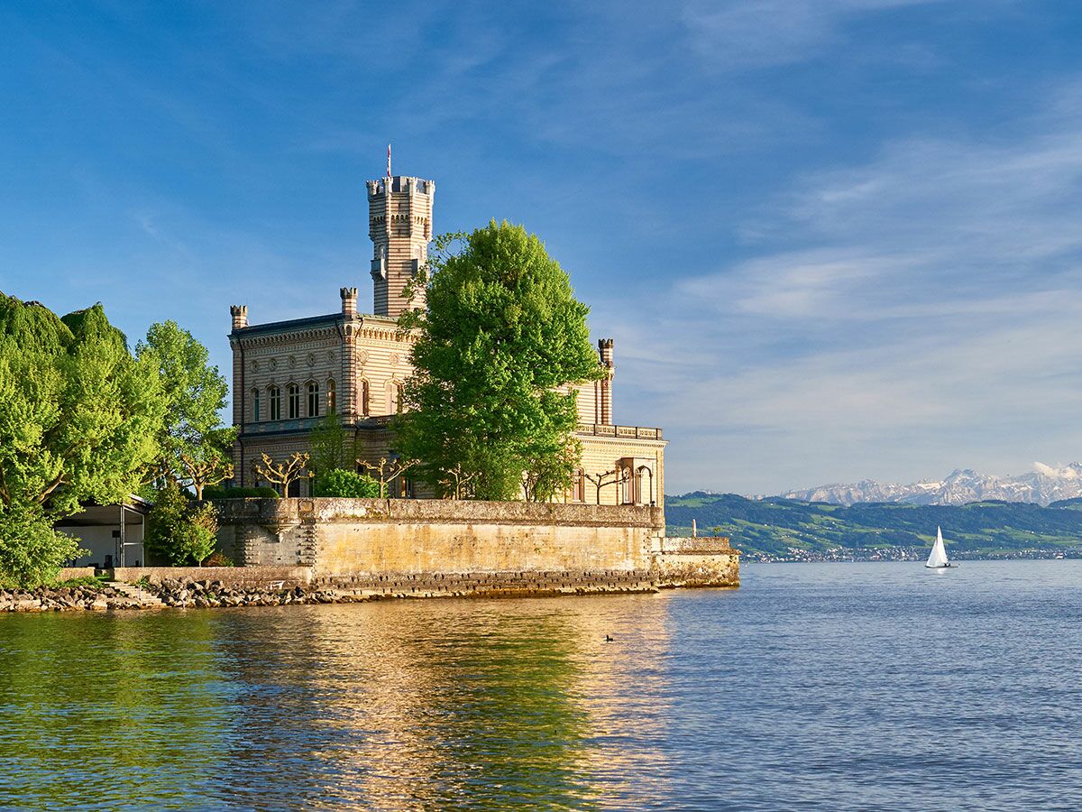 Historisches Schloss mit Turm direkt am Zürichsee unter blauem Himmel mit Segelboot und Alpen im Hintergrund.