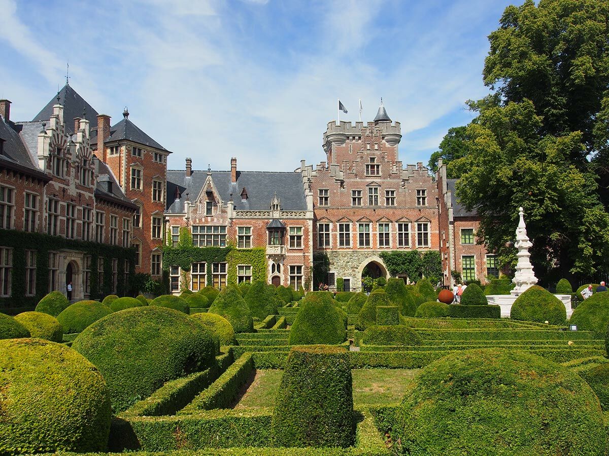 Blick auf ein historisches Backsteinschloss mit Türmen und einem weitläufigen Formschnittgarten unter blauem Himmel.
