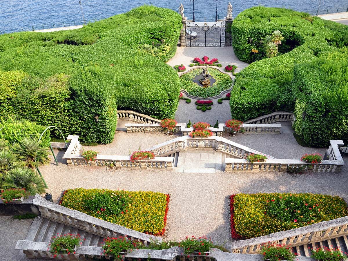 Malerischer terrassierter Garten mit Buchsbaumhecken, Brunnen, Stein-Treppen und Blumenbeeten am See.