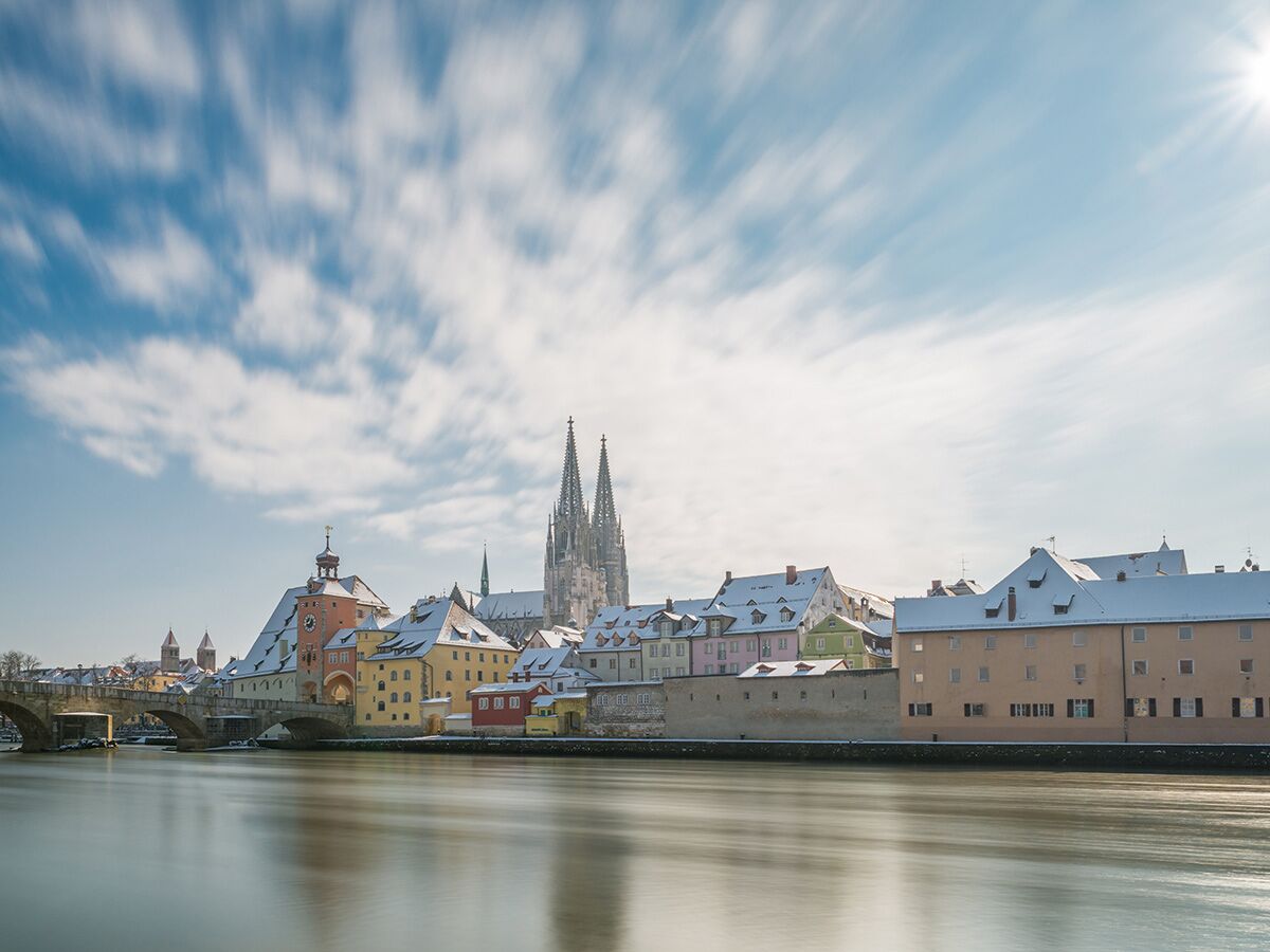 Panoramablick auf Regensburg im Winter mit schneebedeckten Dächern, Steinerner Brücke, Dom St. Peter und Donau bei Sonnenlicht.