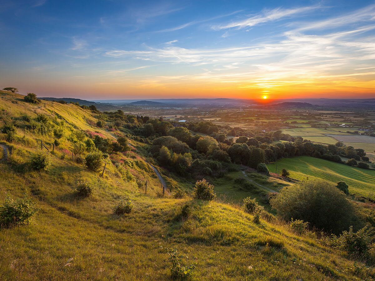 Malerischer Sonnenuntergang über einer weiten Hügellandschaft und einem Tal mit Feldern und Wegen bei goldenem Licht.
