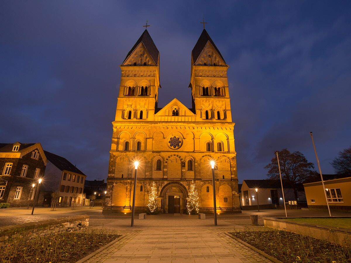Eindrucksvolle romanische Kirche mit zwei Türmen bei Nacht festlich beleuchtet, mit Weihnachtsbäumen und Straßenlaternen auf dem Vorplatz.