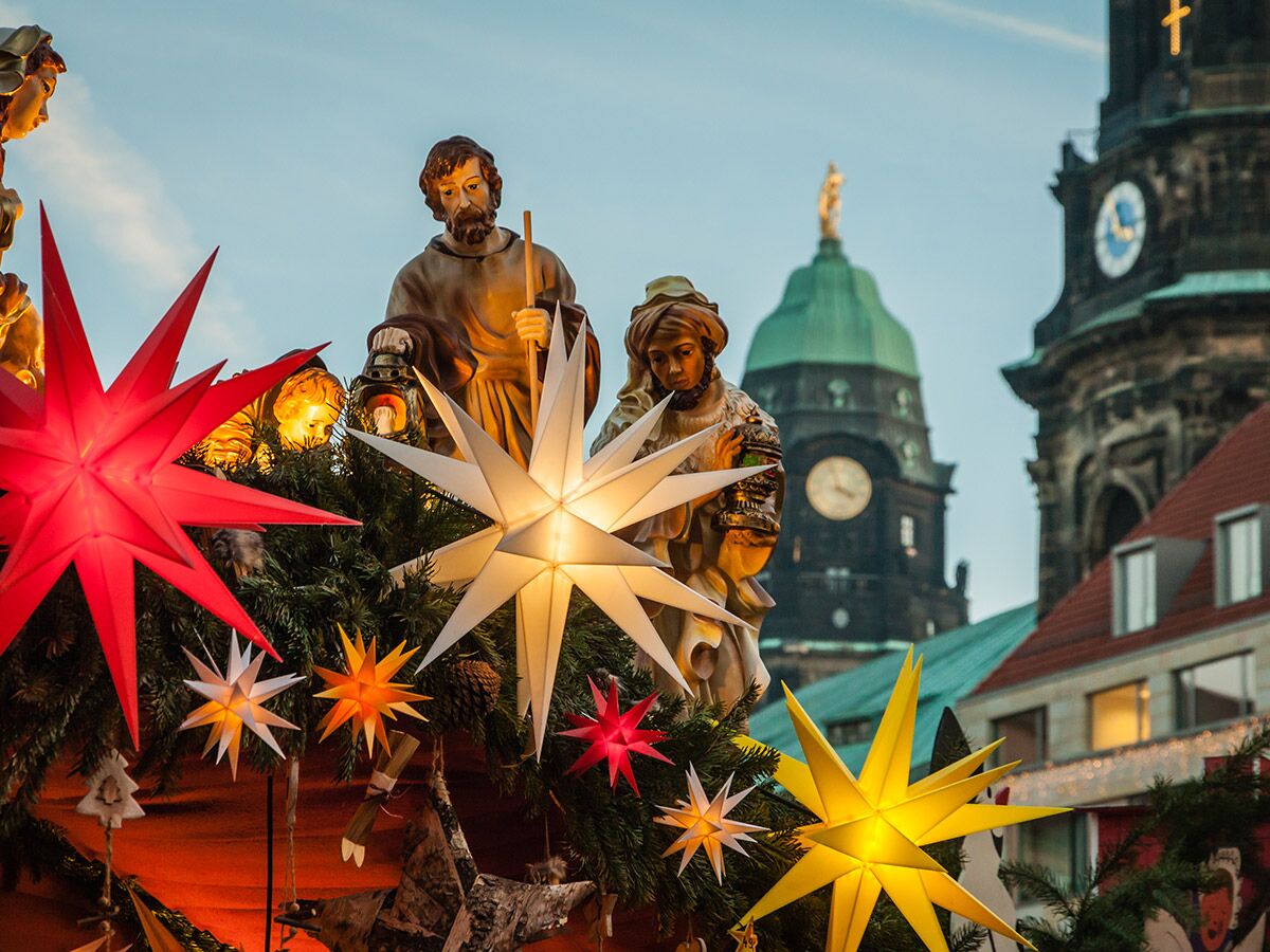 Detailansicht eines Weihnachtsmarktes mit beleuchteten Herrnhuter Sternen, Krippenfiguren und Kirchtürmen im Hintergrund.
