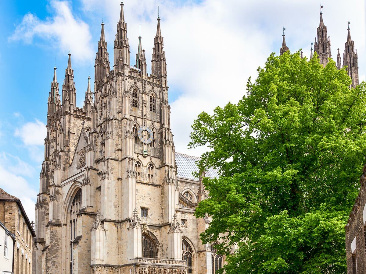 Majestätische Canterbury Kathedrale mit ihren gotischen Türmen und einem großen grünen Baum unter blauem Himmel.