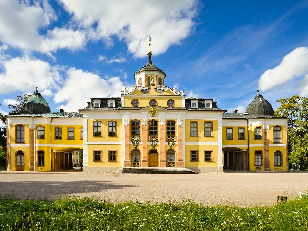 Gelbes Barockschloss mit Kuppeln und zentralem Turm unter blauem Himmel mit weißen Wolken, umgeben von Grünflächen.