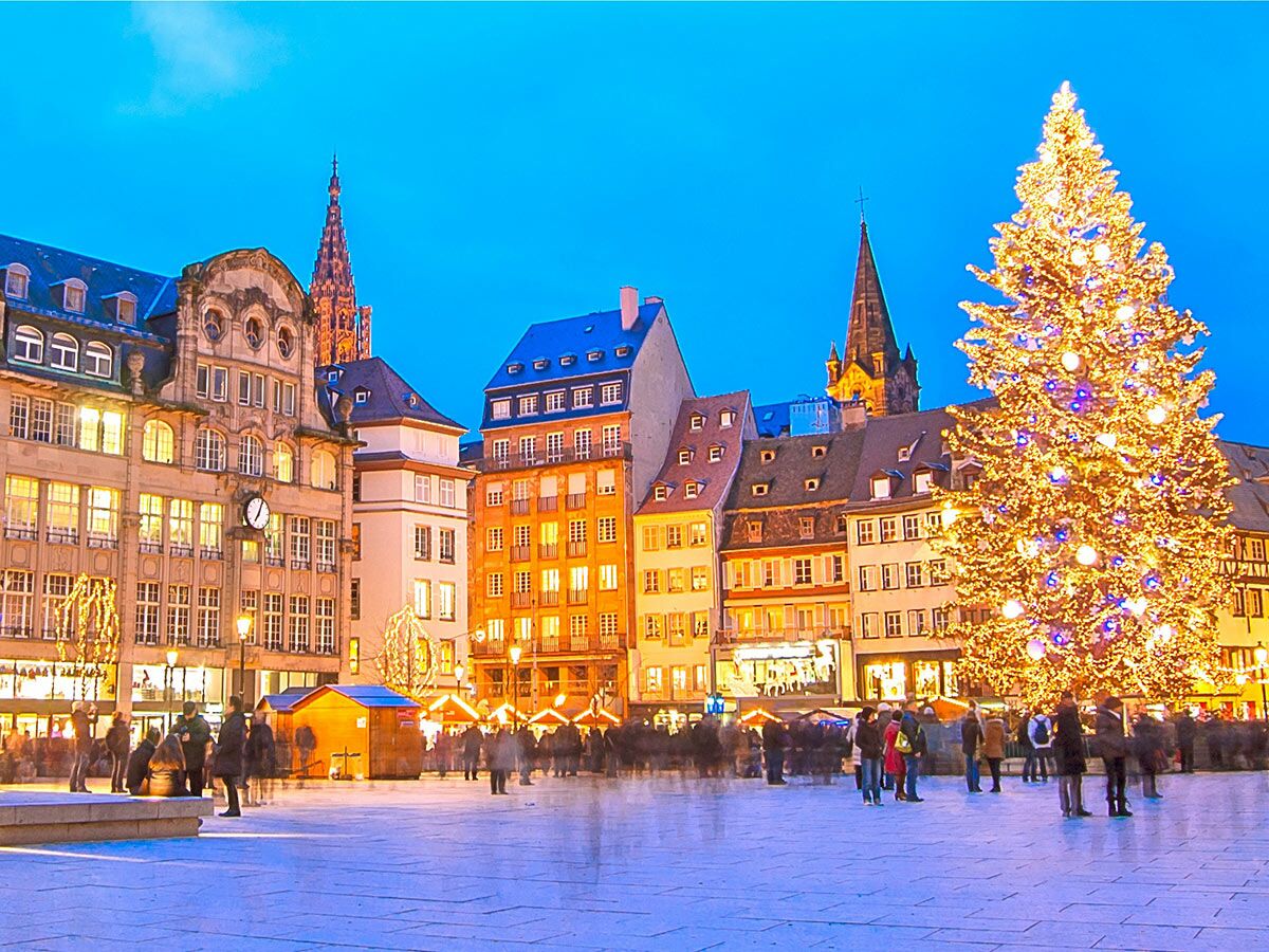 Festlicher Weihnachtsmarkt in Straßburg am Abend mit großem leuchtendem Weihnachtsbaum, historischen Gebäuden und vielen Menschen.