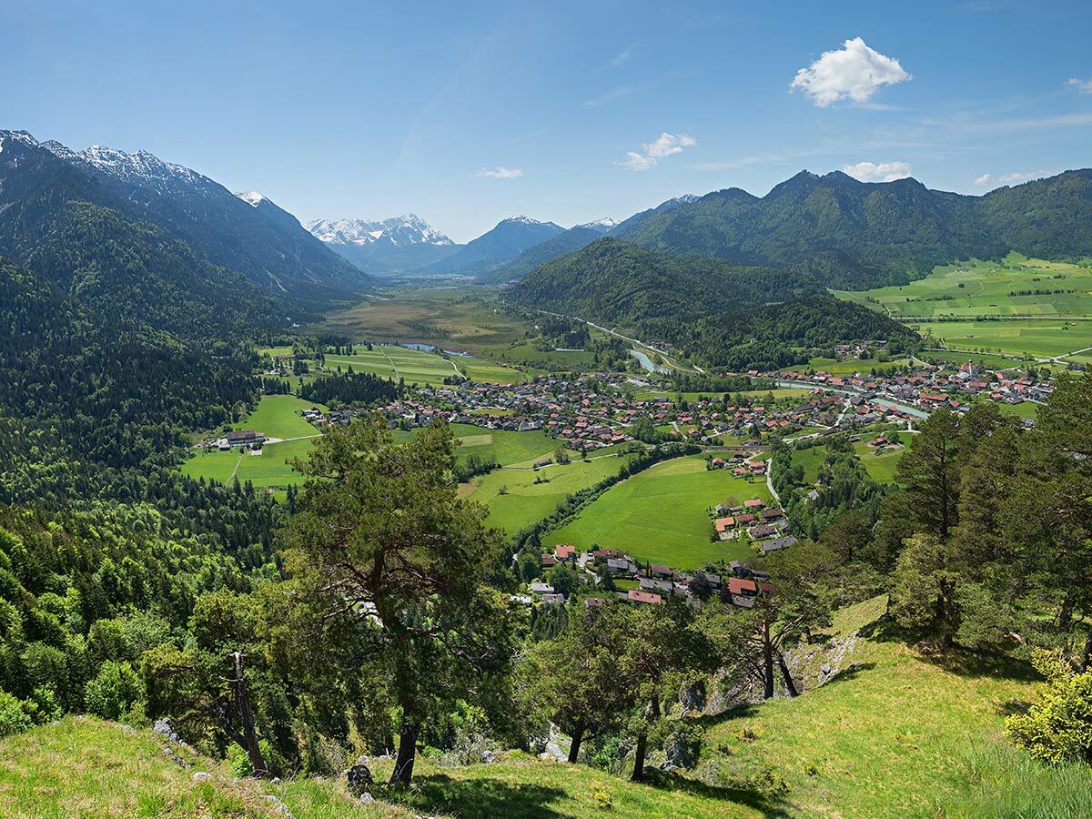Panoramablick auf ein Dorf im grünen Alpetal, umgeben von bewaldeten Bergen und schneebedeckten Gipfeln unter blauem Himmel.