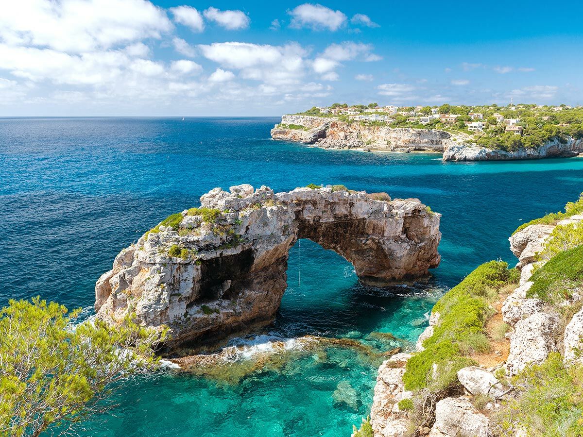 Malerischer Natursteinbogen im türkisen Meer vor einer felsigen Küste mit grüner Vegetation und Häusern unter blauem Himmel.