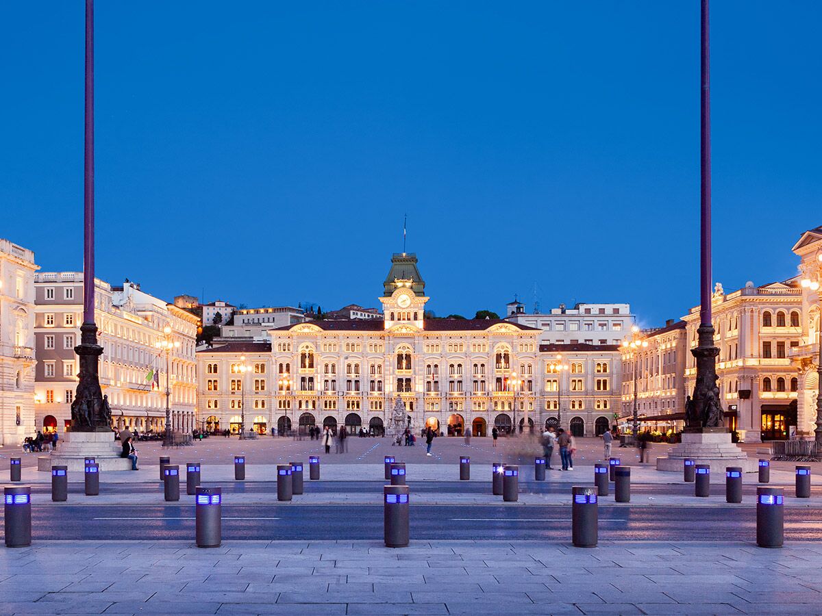 Panoramablick auf die beleuchtete Piazza Unità d'Italia in Triest bei Sonnenuntergang, mit historischen Gebäuden und blauem Himmel.