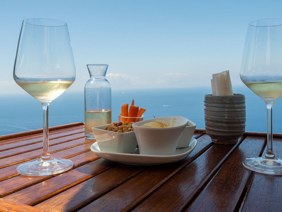 Zwei Gläser Weißwein und Snacks auf einem Holztisch mit atemberaubendem Meerblick unter blauem Himmel.