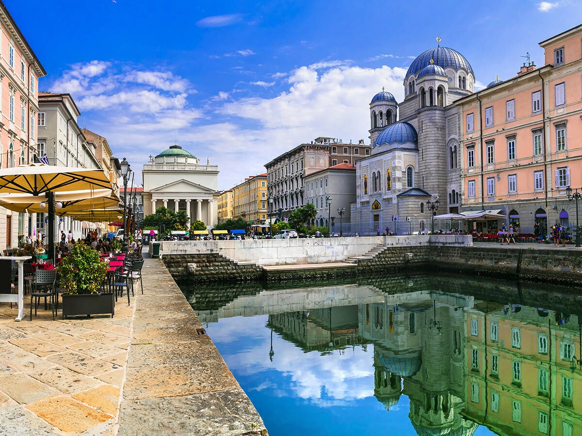 Sonnenverwöhnte Stadtansicht von Triest mit Canal Grande, beeindruckenden Kirchen und Gebäuden, die sich im Wasser spiegeln, und belebten Straßencafés.