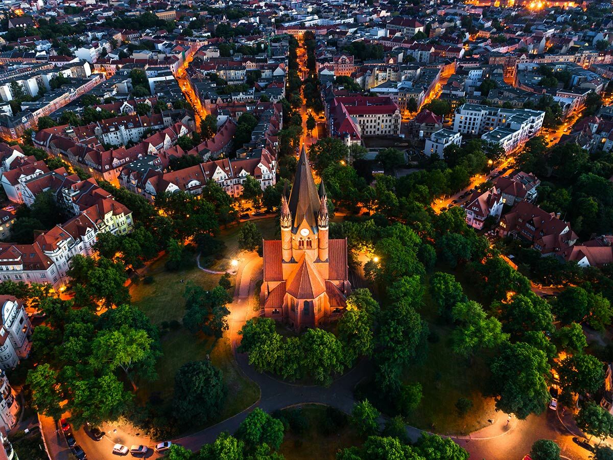 Luftbild einer beleuchteten Kirche und eines Parks bei Nacht, umgeben von städtischen Gebäuden mit roten Dächern.
