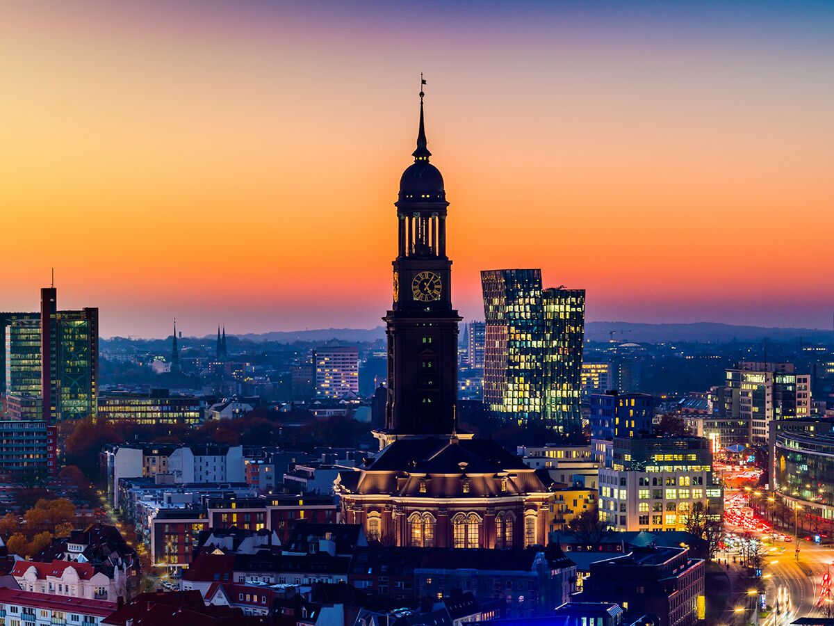 Panoramablick auf Hamburg bei Sonnenuntergang mit der beleuchteten St. Michaelis Kirche und modernen Gebäuden.