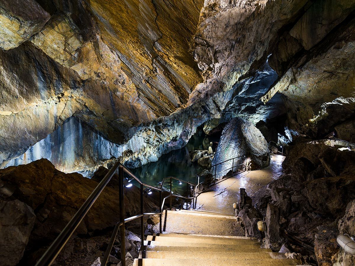 Beleuchtete Höhle mit beeindruckenden Felsformationen, einem unterirdischen See und einem gut ausgebauten Weg mit Geländer.